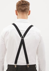 Tirantes de tela negra con clips de metal, que cuentan con un conector triangular, usados sobre una camisa blanca, combinados con pantalones de cuadros grises.