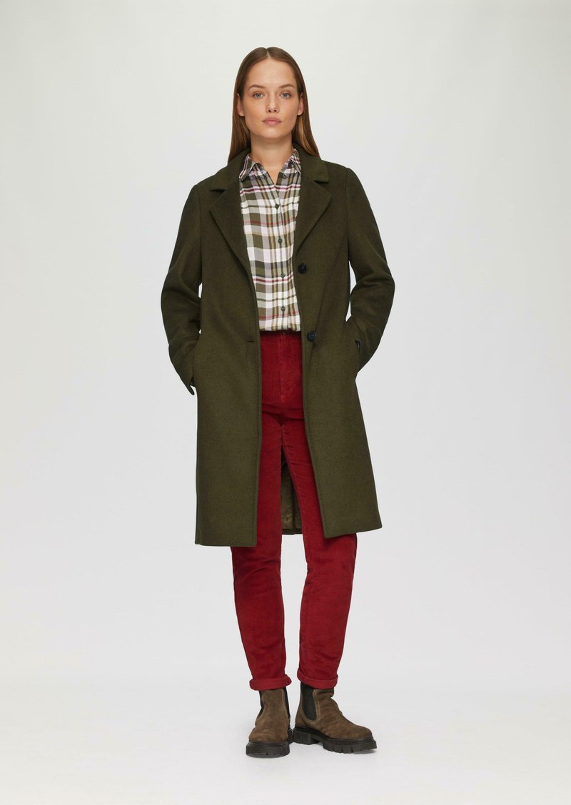 Olifgroen wollen jas met een gelapte kraag, over een geruite blouse. Rode corduroybroek en bruine enkellaarsjes met zwarte zolen maken de outfit compleet.