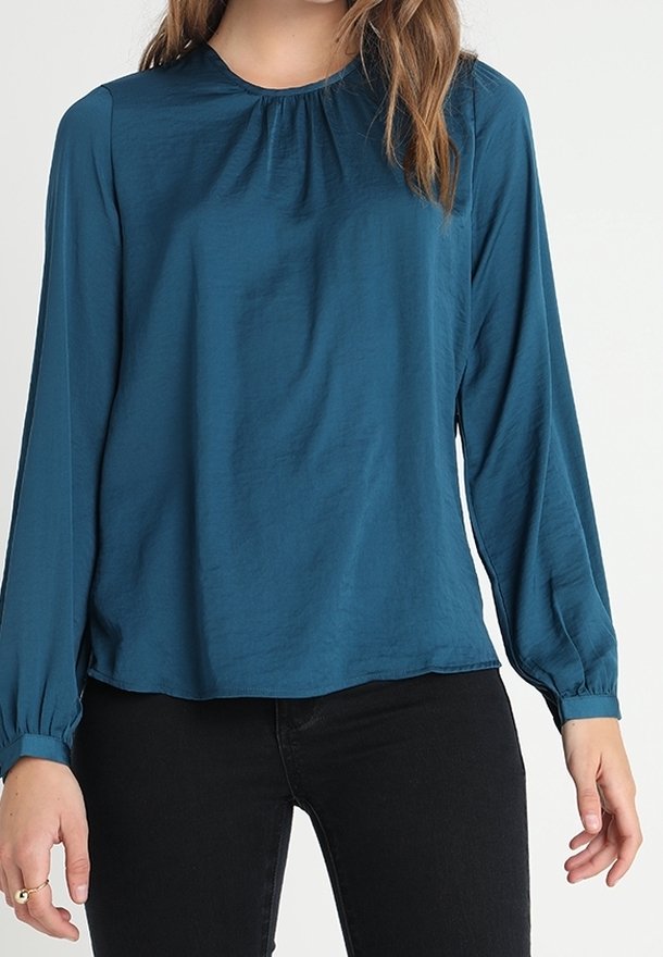 Femme portant un chemisier à manches longues turquoise avec un col froncé et des poignets assortis, associé à un pantalon noir, sur un fond uni.