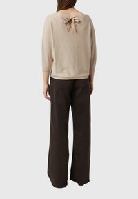 Beżowy sweter dziany z detalem na plecach w formie klucza, związanym jedwabną wstążką, w połączeniu z brązowymi spodniami o szerokich nogawkach. Miękka faktura, luźny krój.
