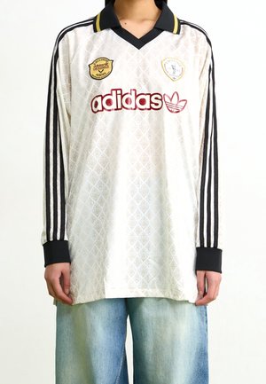 Osoba nosząca białą, vintage, długorękawową koszulkę Adidas z czarnymi paskami, czarnym kołnierzem i logo, w połączeniu z jasnoniebieskimi dżinsami.