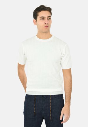 Giovane uomo con capelli corti e scuri che indossa un maglione bianco a maniche corte con trama e jeans blu scuro con coulisse, che guarda di lato.