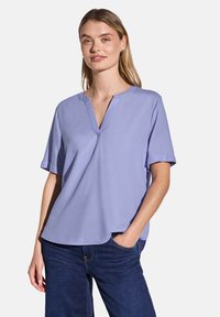 Blusa corta a maniche corte in lavanda con scollo a V, texture liscia, vestibilità rilassata e orlo leggermente asimmetrico, abbinata a jeans scuri.