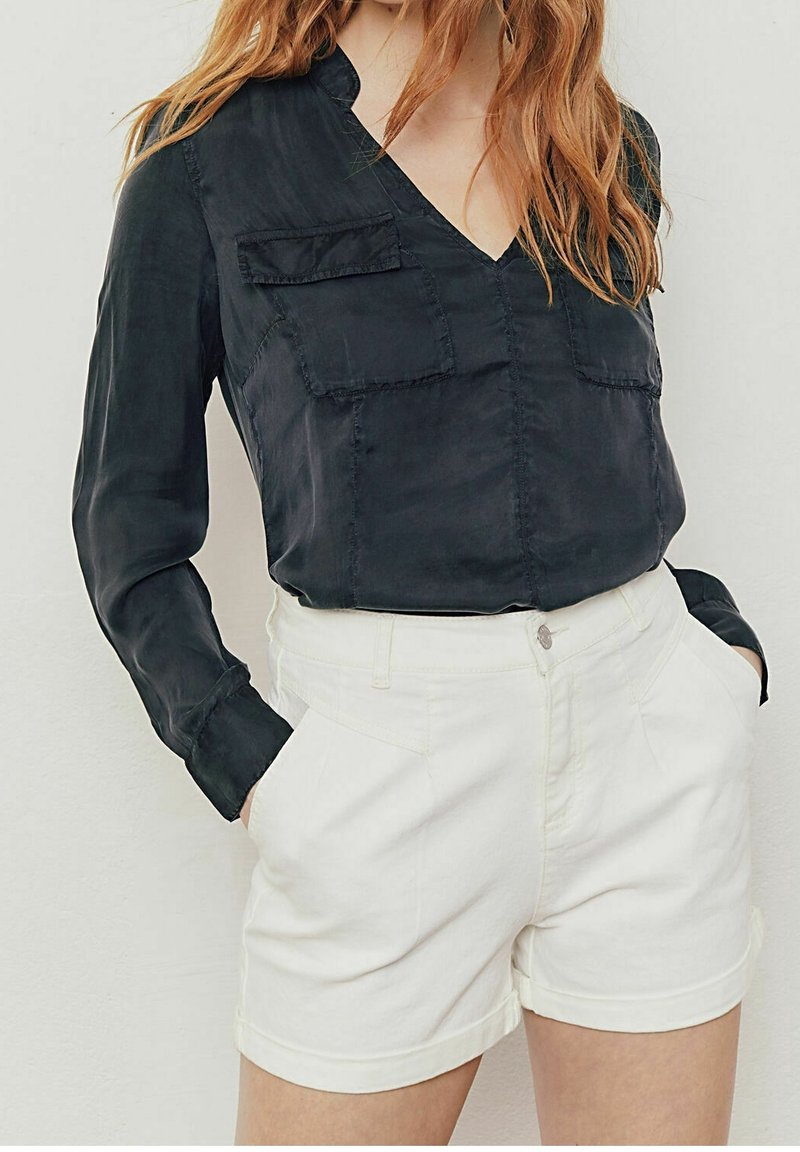 Femme portant un chemisier noir à manches longues avec deux poches poitrine associé à un short blanc taille haute, mains dans les poches, sur fond uni.