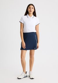 Camisa polo blanca con detalles negros, combinada con una falda corta azul marino con ribetes blancos y zapatillas blancas con acentos oscuros.