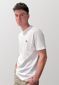 T-shirt bianca in cotone con scollatura rotonda, con un piccolo motivo ricamato sul petto. Modello che indossa pantaloni corti beige casual.