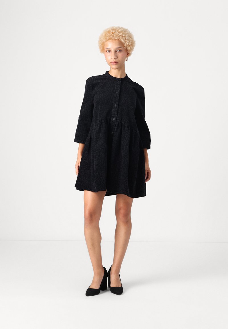 Melna, teksturēta kleita ar pogu priekšpusi, apaļu apkakli un trīs ceturtdaļu piedurknēm. Izplešamas dizains ar iekrāsotu jostasvietu. Valkāta ar melniem papēžiem.