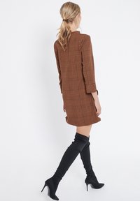 Brun rutig miniklänning med hög halsringning och långa ärmar, i kombination med svarta högklackade stövlar som går upp till låret. Slät tygkvalitet, välstrukturerad passform.