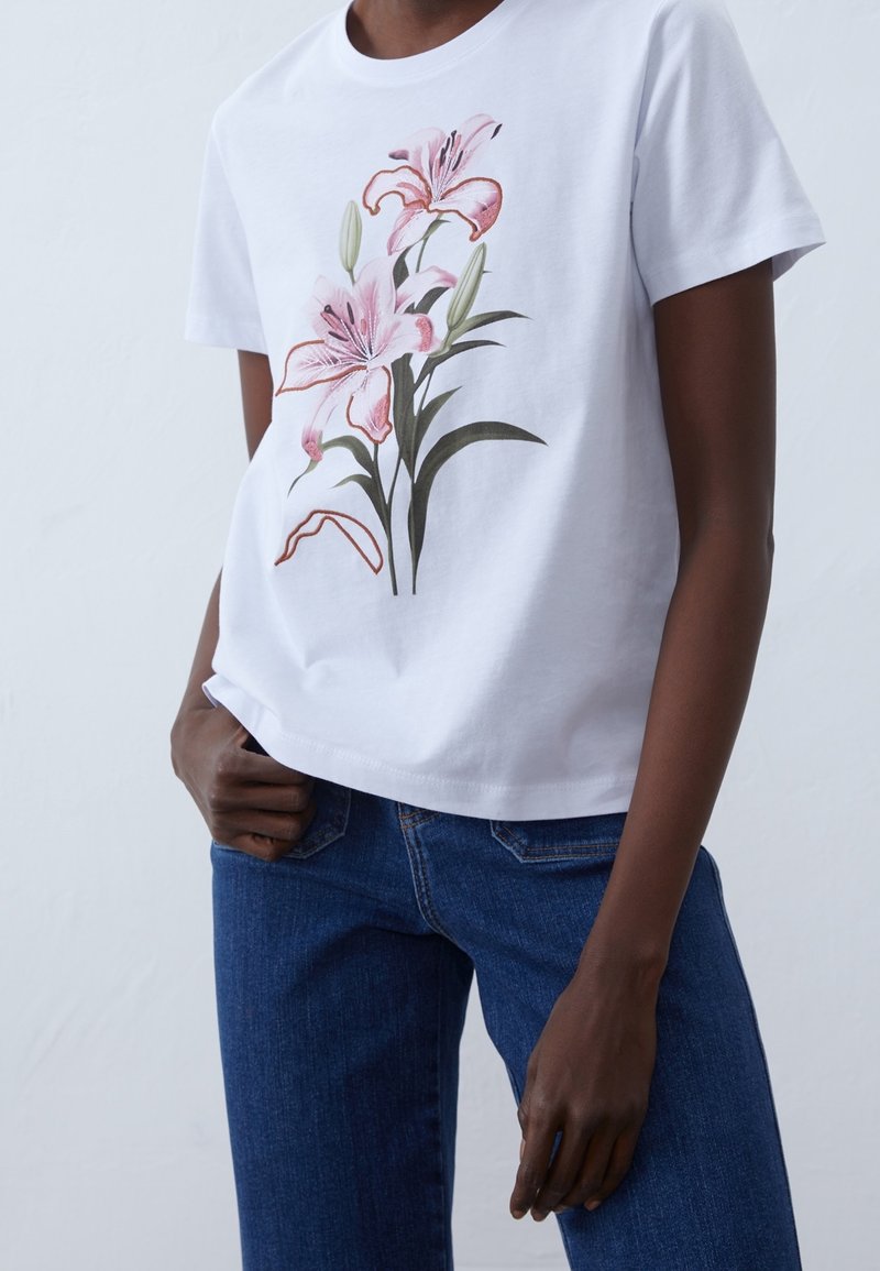 T-shirt en coton blanc avec un motif floral de lys roses et des feuilles vertes, col rond et manches courtes, porté avec un jean bleu.