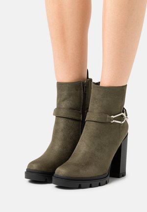Stivaletti alla caviglia in suede verde oliva con tacco spesso nero, chiusura con zip e una fascia decorativa con un accento in metallo argentato.