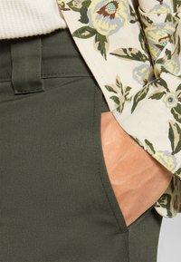 Olive green, tapered trousers with side pockets; cotton fabric with a smooth texture. A floral, cream-colored jacket partially visible.