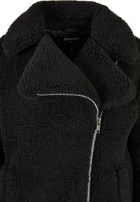 Sort jakke i imiteret pels med sjalskrave, fuld lynlås og struktureret stof. Har en sidelomme med lynlås og et blødt, fluffy ydre.