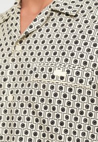 Korte mouw shirt in crèmekleurige stof met een zwart geometrisch patroon, knoopsluiting en een opgestikte zak met stikseldetails.