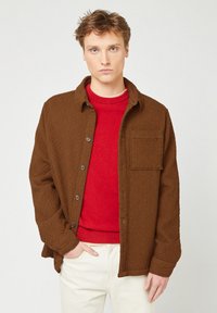 Junger Mann trägt eine texturierte braune Knopfjacket über einem roten Pullover, mit einer Hand in der Tasche, helle Hosen, neutraler Hintergrund.