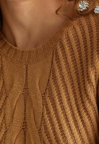 Zbliżenie na osobę ubraną w brązowy sweter w warkoczowy splot z skręconymi wzorami oraz srebrne kolczyki ozdobione kamieniami.