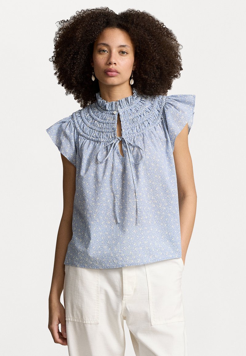 Lichtblauwe bloemenblouse met ruches aan de halslijn en korte mouwen. Heeft een sleutelgat voorkant met strik en een subtiel wit bloemenpatroon.