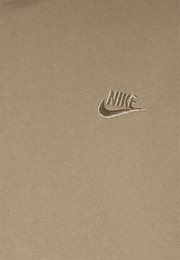 Tan ribbad tyg t-shirt med en subtilt broderad vit Nike-logga på vänster bröstområde. Slät textur, avslappnad design.