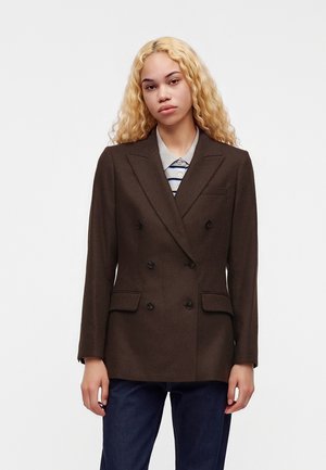 Jeune femme aux longs cheveux blonds bouclés porte un blazer marron à double boutonnage sur une chemise rayée à col et un jean bleu foncé.