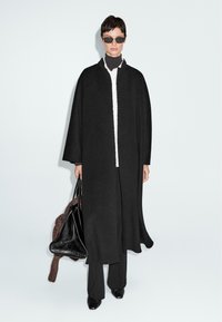 Persona con capelli corti e scuri, che indossa un ampio cappotto nero, una camicia bianca, pantaloni neri, occhiali da sole, mentre tiene una grande borsa nera su uno sfondo chiaro.