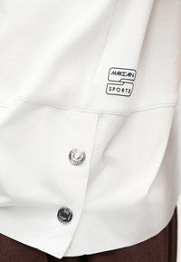 Λευκό ένδυμα Marc Cain Sports με ραμμένο λογότυπο και δύο μεταλλικά κουμπιά τύπου snap με χάραξη το όνομα της μάρκας.