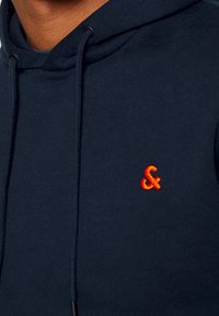 Mörkblå sweatshirt med huva, prydd med en orange broderad logotyp på bröstet. Slät yta med justerbara snören.
