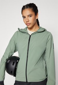 Leichte, strukturierte grüne Jacke mit Reißverschluss und Kapuze. Mit Seitentaschen und einem Logo-Patch, kombiniert mit einem schwarzen Helm.