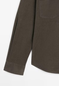 Długi rękaw ciemnobrązowej koszuli z mankietem i widocznym przeszyciem krawędzi kieszeni na białym tle.