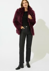 Cappotto in pelliccia sintetica color bordeaux con superficie testurizzata, top nero, pantaloni in pelle nera a vita alta e stivaletti neri con tacco.