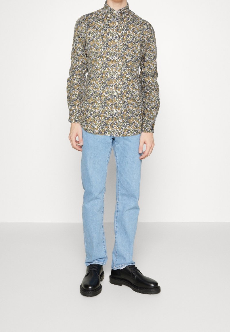 Personne portant un chemisier à manches longues à col montant avec un motif floral, un jean droit bleu clair et des chaussures en cuir noires à lacets.