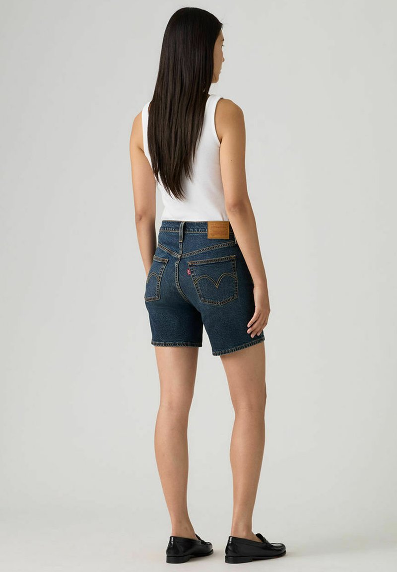 Shorts en denim bleu foncé, taille haute avec ourlet effiloché. Comprend des poches arrière et une écusson en cuir marron à la ceinture. Mocassins noirs portés.