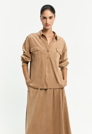 Frau mit zurückgekämmtem Haar, die ein lockeres, beiges Hemd mit Knöpfen und Taschen sowie einen passenden Rock trägt, Hände in den Taschen, neutraler Hintergrund.