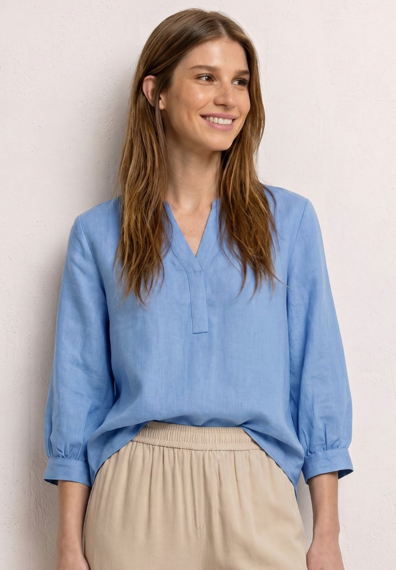 Femme souriante aux longs cheveux bruns porte un chemisier bleu clair à manches trois-quarts et un pantalon beige à taille élastique, debout contre un mur pâle.