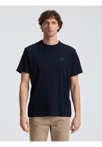 Camiseta de manga corta azul marino con un patrón de rayas texturizadas y un pequeño parche del logo en el lado izquierdo del pecho. Corte casual con cuello redondo.