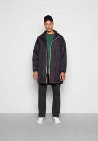 Veste noire imperméable avec une fermeture éclair jaune, capuche ajustable et manches longues sur une chemise verte, associée à un jean foncé et des baskets blanches.