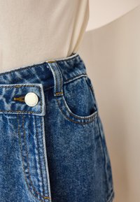 Donkere blauwe denimrok met een hoge taille, voorzien van opvallende stiksels, een metalen knoop en zijzakken. Zachte textuur.