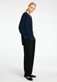 Marineblå langærmet bluse med v-hals, parret med sorte bred benbukser og sorte slip-on sko; glat stof med en underspillet silhuet.