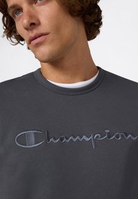 Młody mężczyzna z kręconymi włosami, ubrany w ciemnoszary dres Champion nałożony na biały t-shirt z okrągłym dekoltem, patrzy lekko w górę.