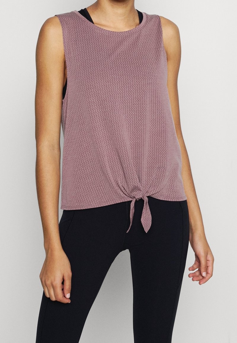 Femme portant un haut texturé mauve sans manches avec un nœud devant, assorti à un legging noir, debout devant un fond clair et uni.