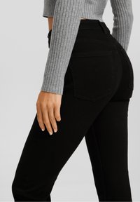 Jeans de mezclilla negra con forma ajustada, cintura alta y dos bolsillos traseros. La mujer lleva una camiseta de manga larga gris con canalé.