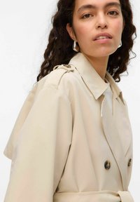 Beige trenchcoat med en mjuk textur, dubbelknäppt design, knappramar och en överdimensionerad krage. Har axelklaffar.
