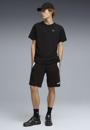 Jonge man kijkt naar links, draagt een zwarte Puma-pet, zwart T-shirt, zwarte korte broek met witte zijstrepen, zwarte sokken en zwarte sneakers.