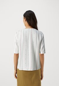 Weiße Bluse mit vertikalen Streifen in Rot, Schwarz und Blau. Kurze Ärmel, runder Halsausschnitt und glatte Textur. Leicht abgerundeter Saum.