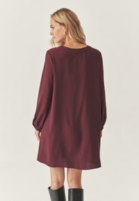 Burgundy pitkähihaine mekko kevyestä kankaasta, jossa on rento istuvuus ja kerättyjä hihansuita, yhdistettynä mustiin polviretkiin saappaisiin.