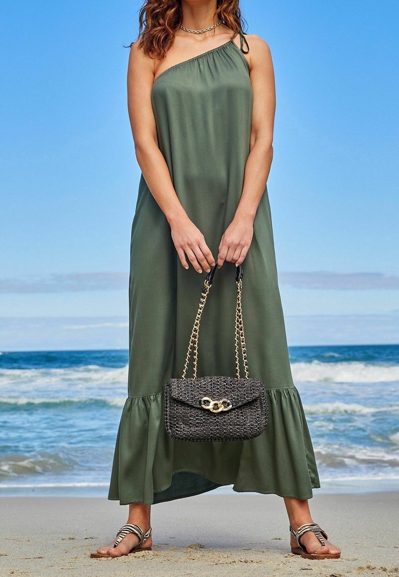 Robe maxi verte à une épaule avec un bas évasé, associée à un sac à main tissé noir avec une chaîne dorée. Portée avec des sandales métalliques.