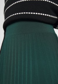 Dunkelgrüner, plissierter Rock mit glatter Textur und breitem Bund, kombiniert mit einem schwarz-weiß gestreiften Pullover oben.