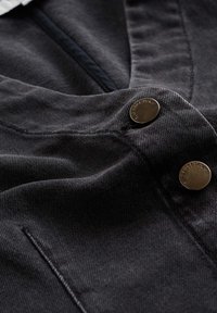 Mörkgrå denimtyg med en smidig textur, framträdande guldmetallknappar och synliga sömnadsdetaljer.