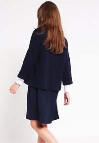 Žena s dlouhými hnědými vlasy, oblečená v námořnickém modrém kostýmku a bílé košili, stojící ráda s opřenými zády a uvolněnými pažemi o bílé pozadí.