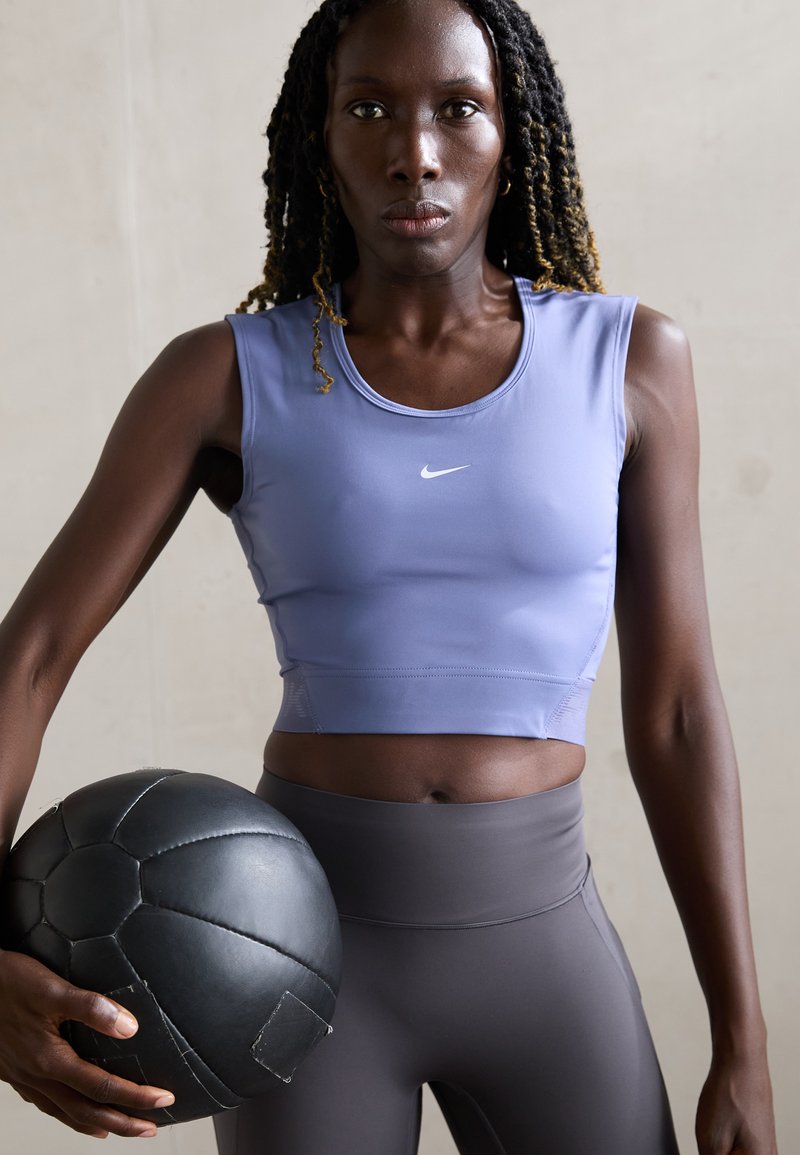 Atletisk kvinde med flettet hår holder en sort medicinbold, iført en lavendelfarvet ærmeløs crop top og grå højtaljede tights.