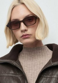 Giacca di pelle marrone con colletto a coste, maglione a coste beige chiaro e occhiali da sole rettangolari con lenti oscurate.