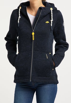 Femme portant un sweat à capuche bleu foncé zippé avec cordons blancs, logo parapluie jaune, et les mains partiellement dans les poches avant, associé à un jean bleu.